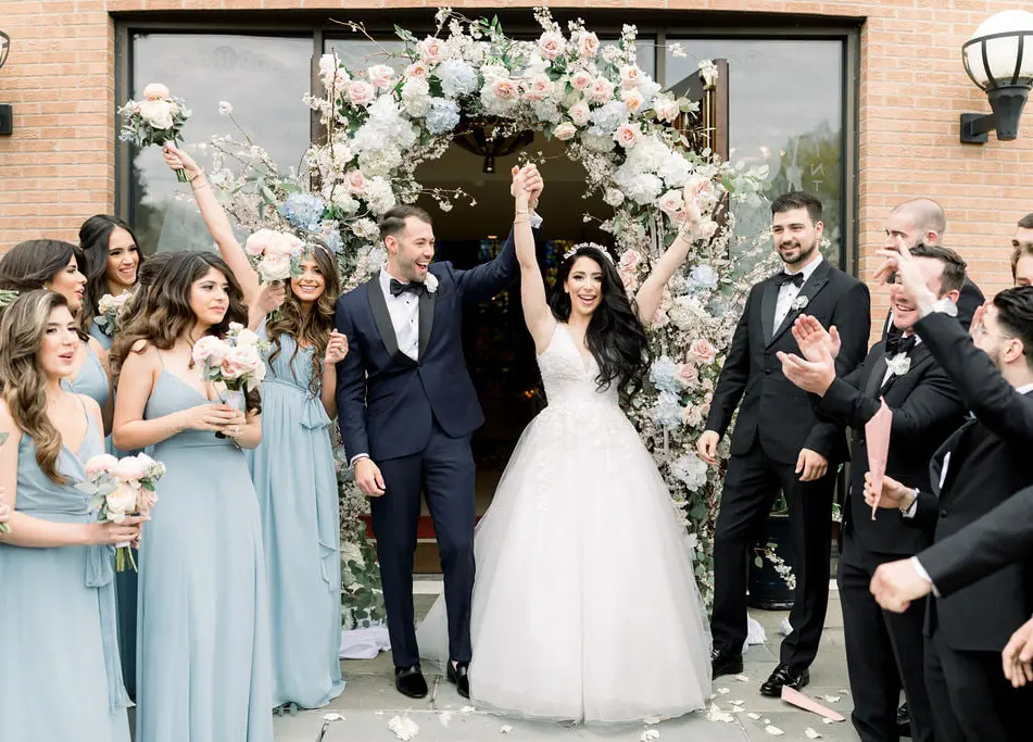 Outside the church, the couple raise their arms beneath an arch of blush and blue flowers as the wedding party cheers and throws petals.