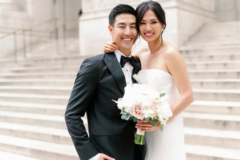 The groom leans in, grinning, beside the bride on the library steps, her bouquet of white and blush roses between them.
