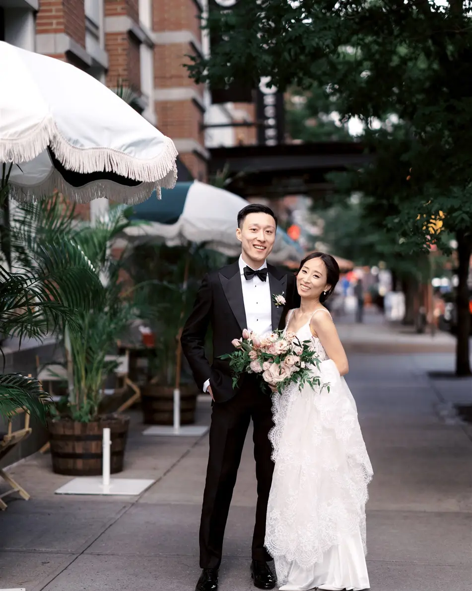 The couple stand on a leafy Tribeca sidewalk beside a white café umbrella, the bride holding a bouquet of pale roses.