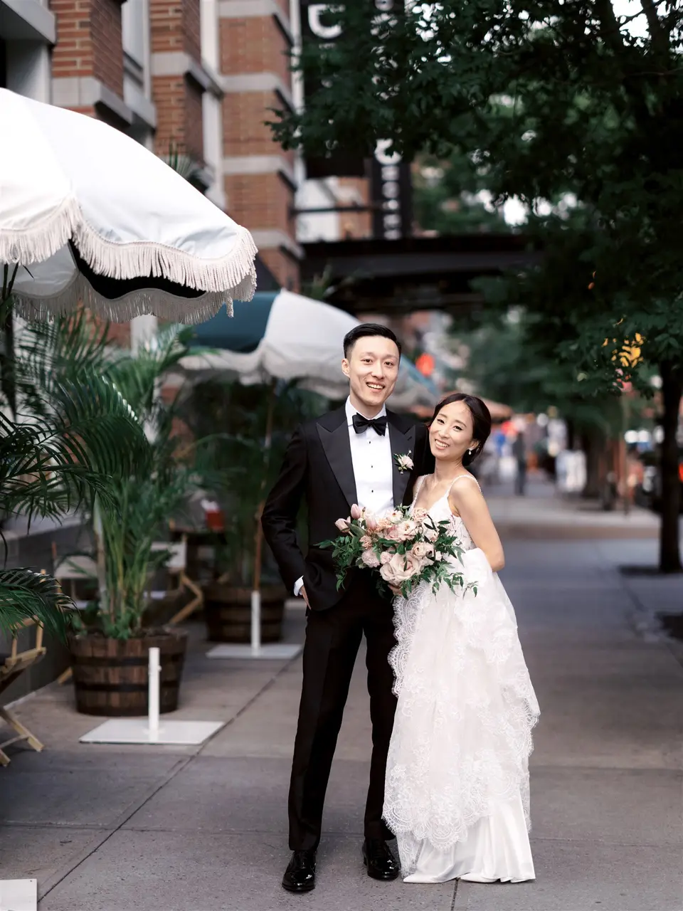 The couple stand on a leafy Tribeca sidewalk beside a white café umbrella, the bride holding a bouquet of pale roses.