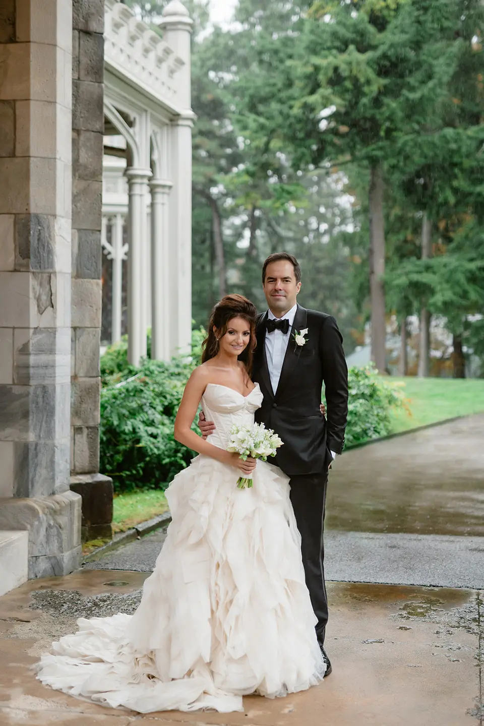 The couple stand beneath the stone arcade of the mansion, the bride's tiered gown spread on the wet path, tall trees behind.