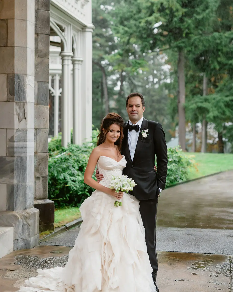 The couple stand beneath the stone arcade of the mansion, the bride's tiered gown spread on the wet path, tall trees behind.