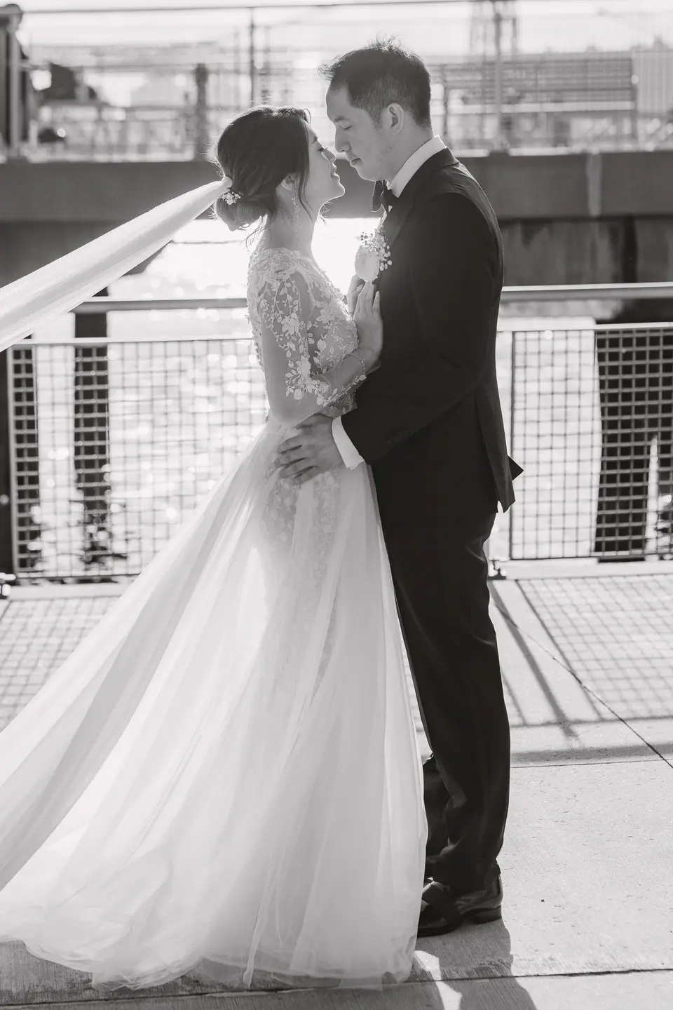 The couple kiss beside a railing on the waterfront, her veil lifting in the wind. Black and white.