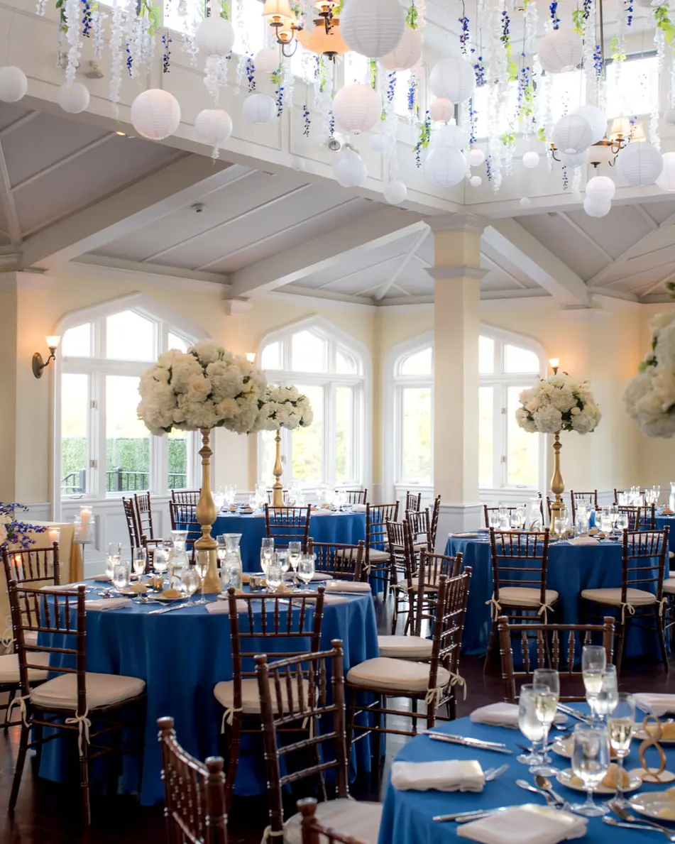 The reception room, round tables in deep blue linen with tall white arrangements beneath hanging paper lanterns.