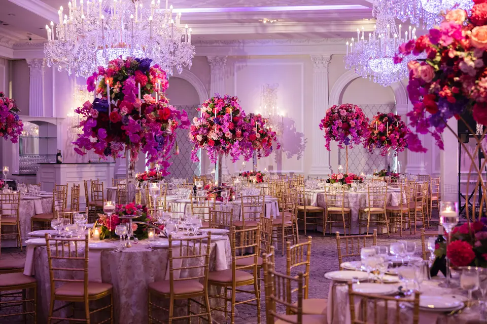 The ballroom set for dinner beneath crystal chandeliers, tall pink and red arrangements above every table.