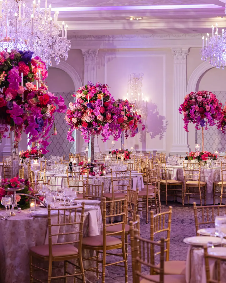 The ballroom set for dinner beneath crystal chandeliers, tall pink and red arrangements above every table.