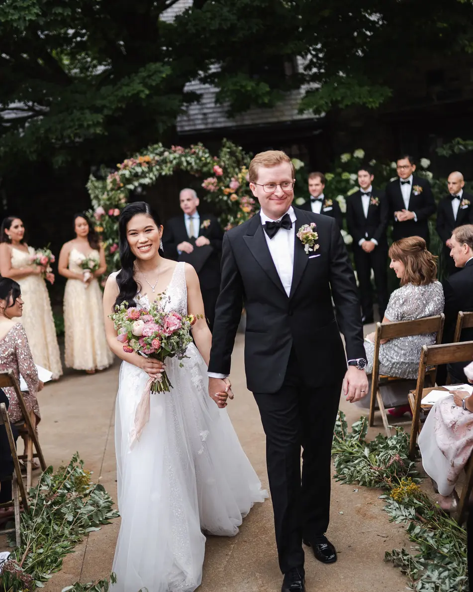 Married, the couple walk back up the aisle between smiling guests, beneath an arch of pink flowers.