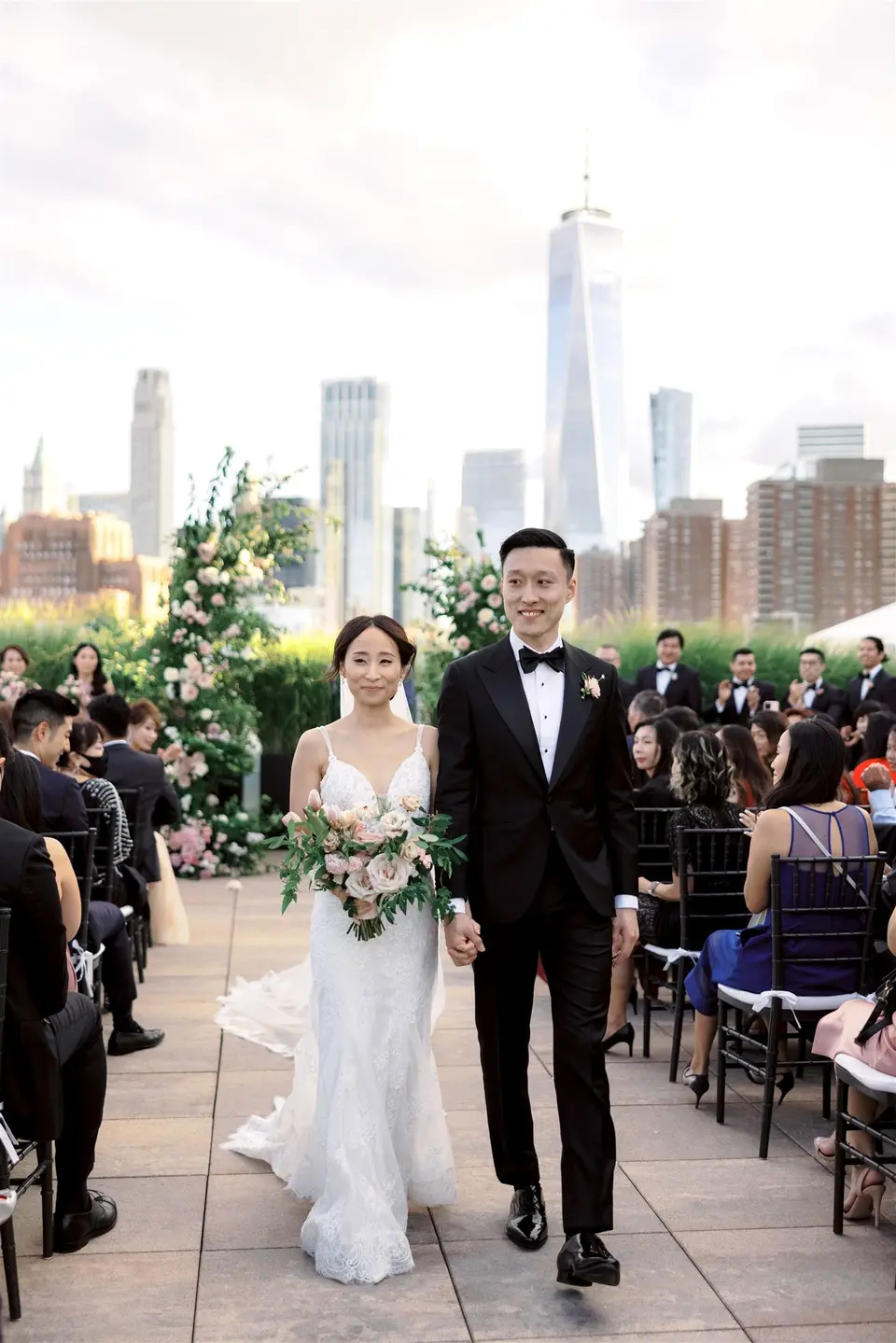 Married, the couple walk back up the aisle hand in hand, guests standing on either side, the towers of Lower Manhattan behind.
