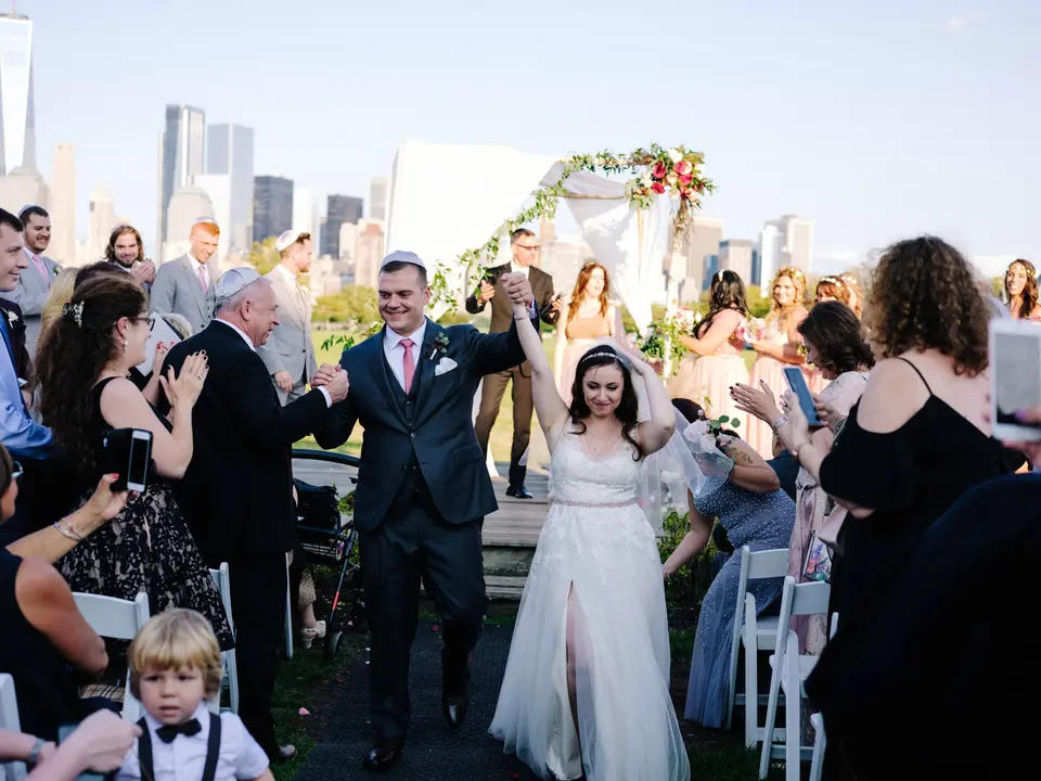 Married, the couple walk back up the aisle to cheering guests, arms raised, the skyline behind them.