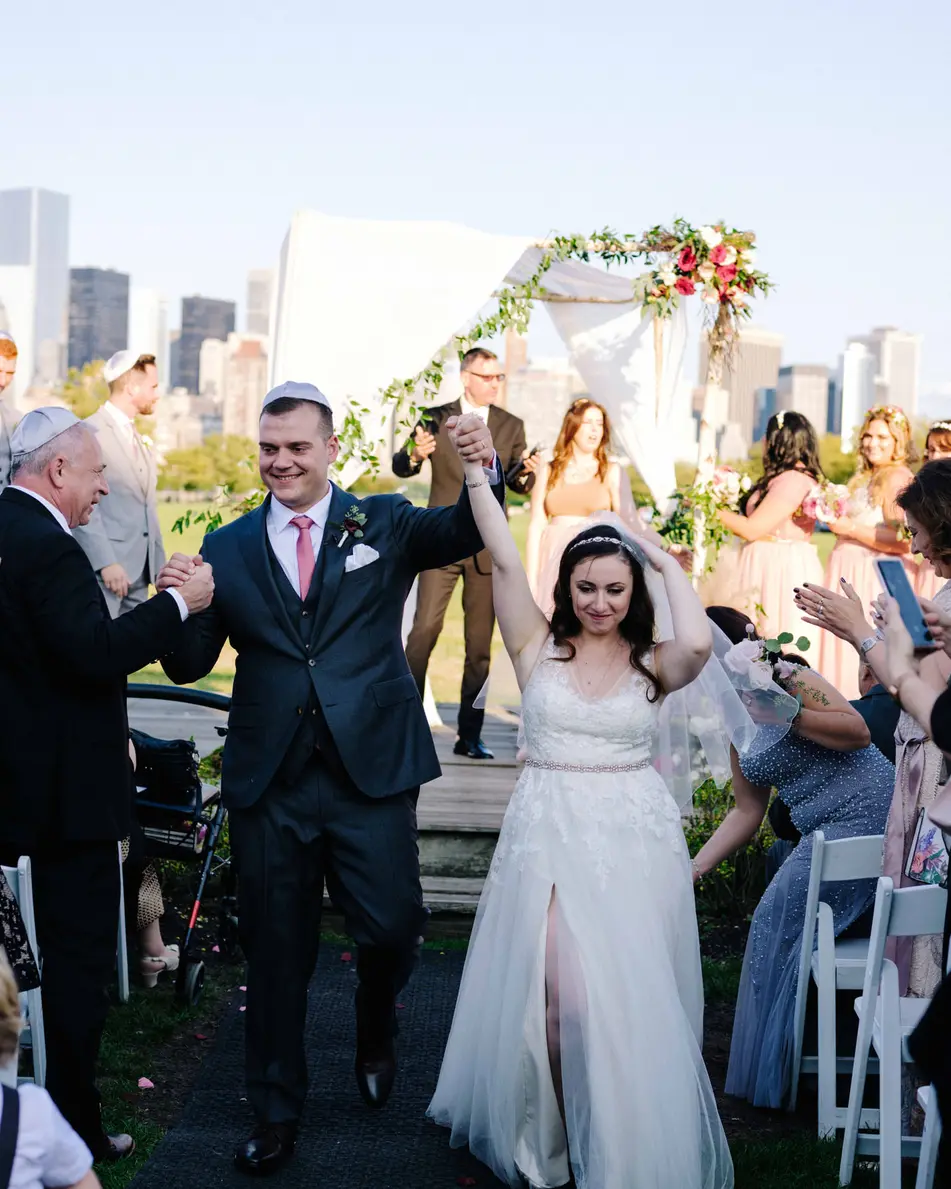 Married, the couple walk back up the aisle to cheering guests, arms raised, the skyline behind them.