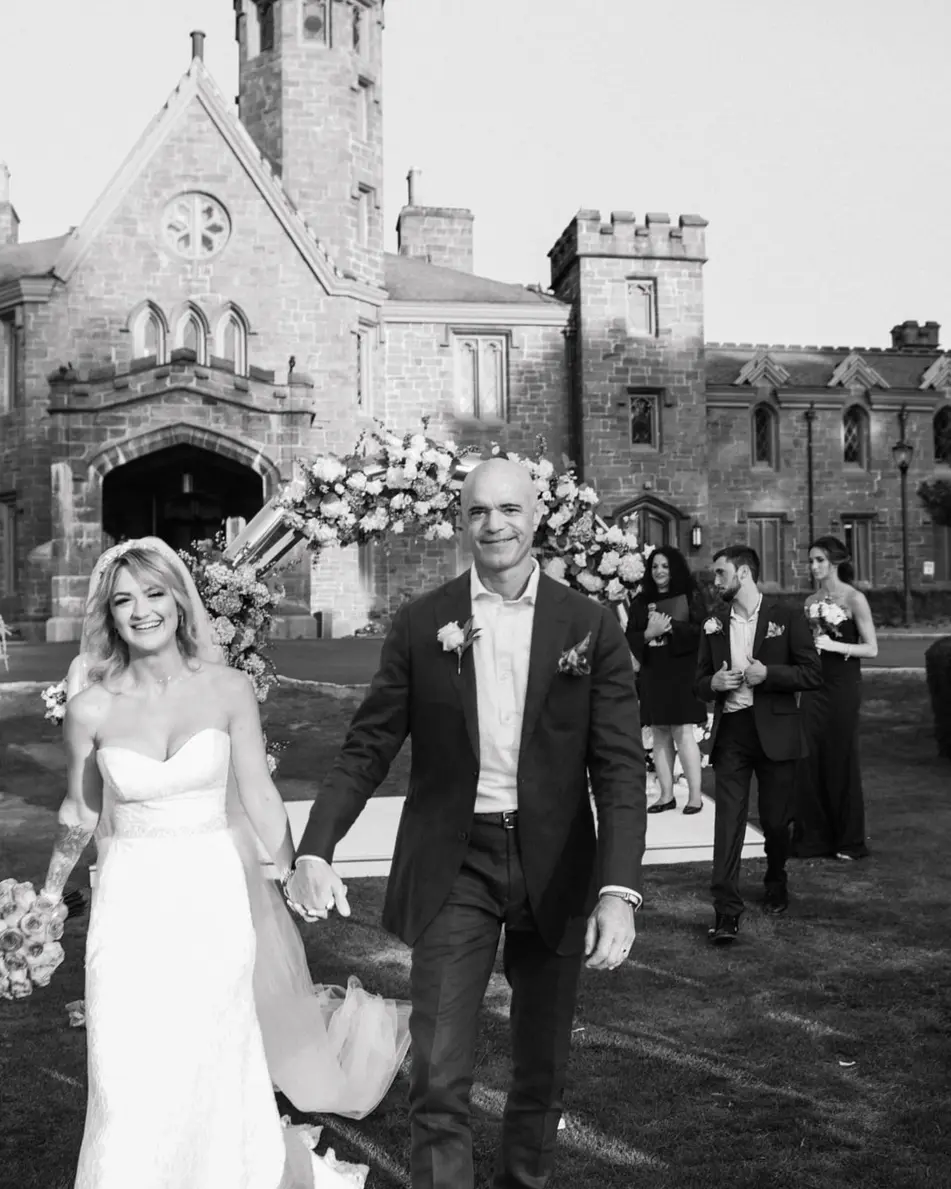 Married, the couple walk back up the aisle between cheering guests, the castle behind them. Black and white.