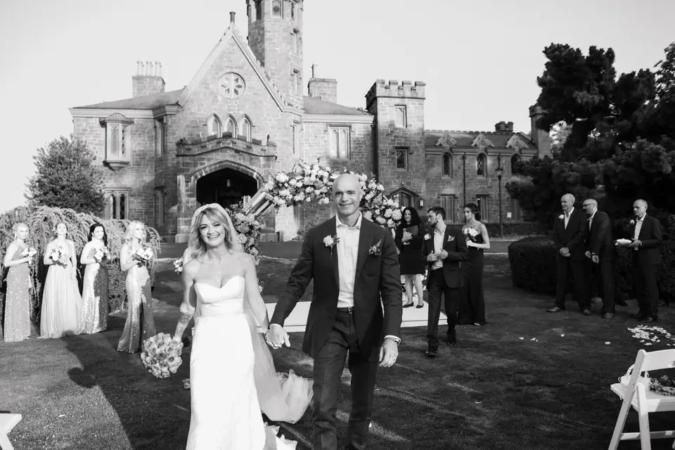 Married, the couple walk back up the aisle between cheering guests, the castle behind them. Black and white.