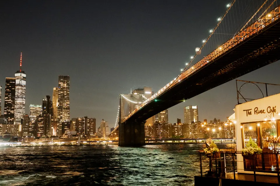 The River Café glowing at the water's edge at night, the Brooklyn Bridge lit above it and Lower Manhattan across the river.