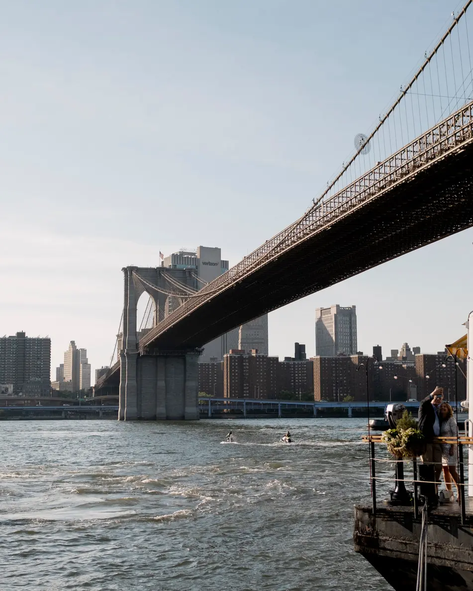 The River Café beneath the Brooklyn Bridge on a bright afternoon, Lower Manhattan across the water.