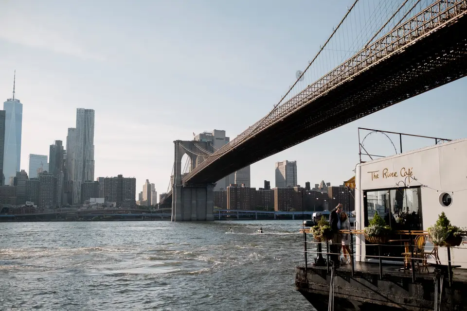 The River Café beneath the Brooklyn Bridge on a bright afternoon, Lower Manhattan across the water.