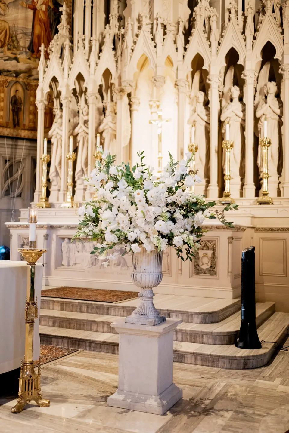A marble altar of carved Gothic spires, an urn of white flowers on a pedestal before it.