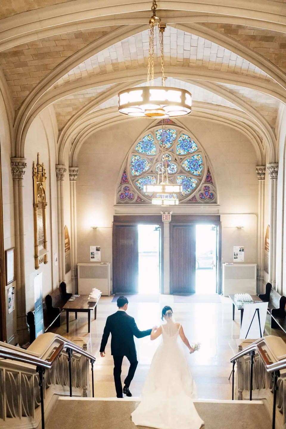 The bride and groom walk down the stairs toward the church doors beneath a rose window.
