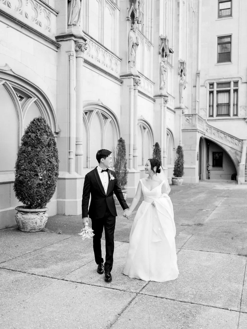 The bride and groom walk hand in hand through a stone courtyard. Black and white.