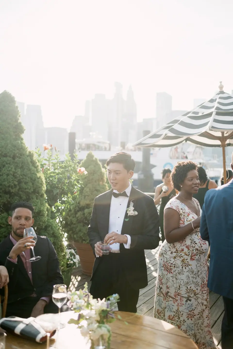 The groom talks with guests at cocktail hour, a striped umbrella and the Lower Manhattan skyline behind.