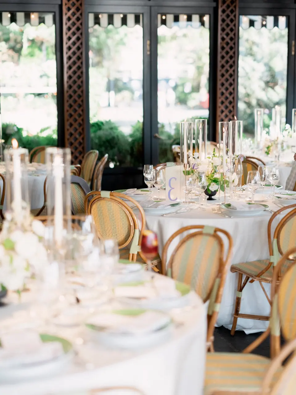 Round tables with rattan chairs and tall glass candle holders by the windows.