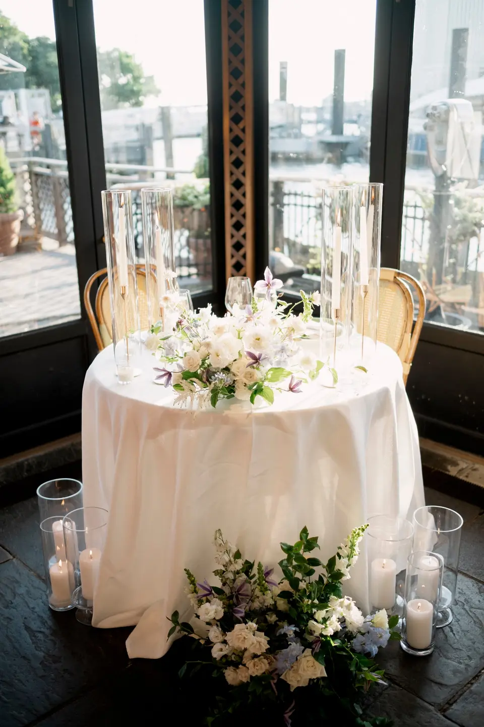 The sweetheart table by the window, white and lavender flowers and tall glass candles around it.