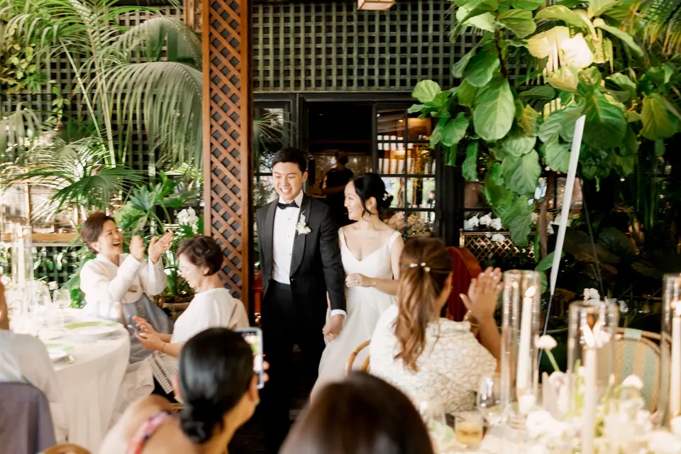 Guests applaud as the bride and groom enter the dining room among palms and candlelit tables.