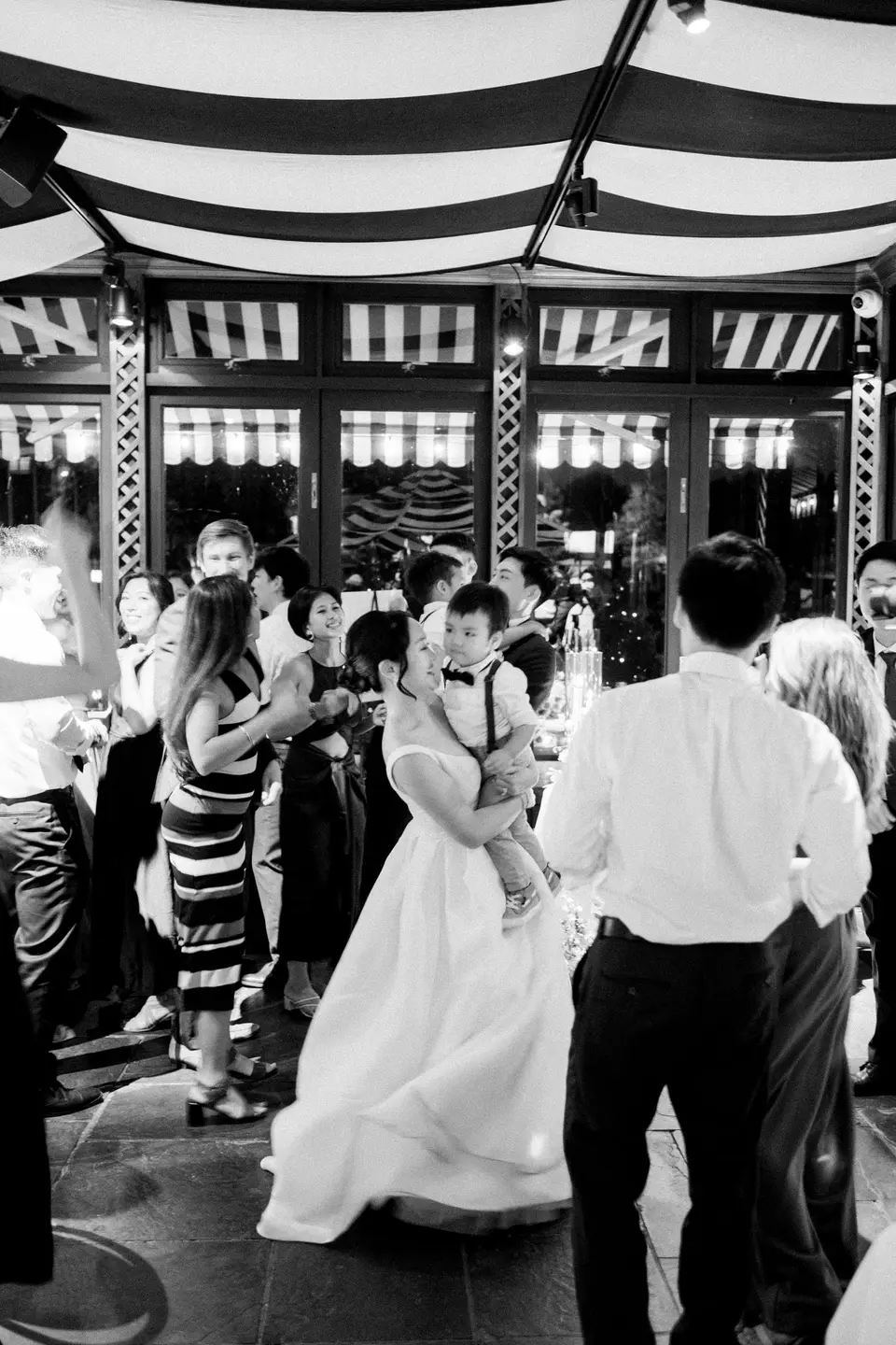The bride dances with guests beneath the striped awning ceiling. Black and white.