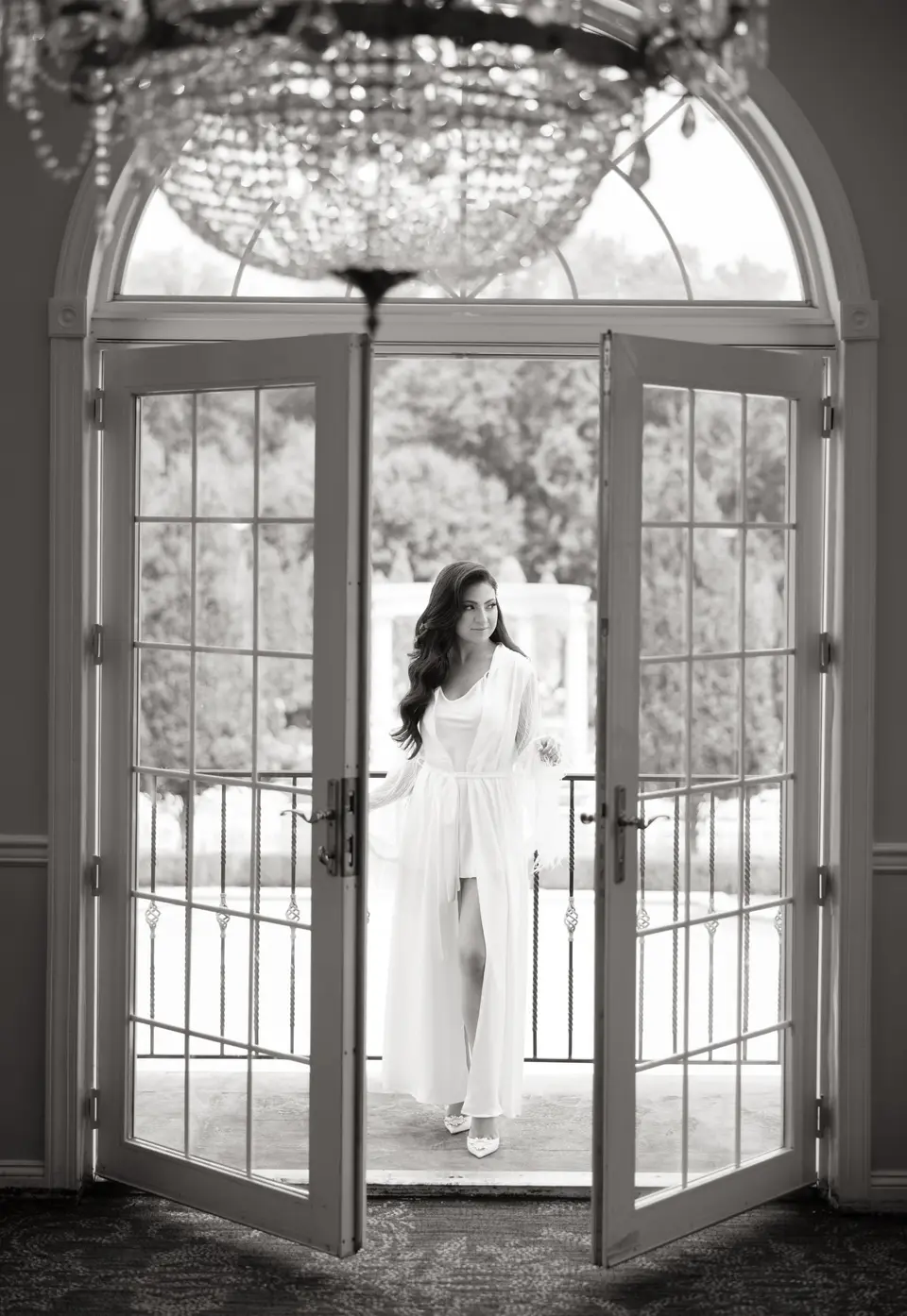 The bride in a robe stands in an open French door beneath a crystal chandelier. Black and white.