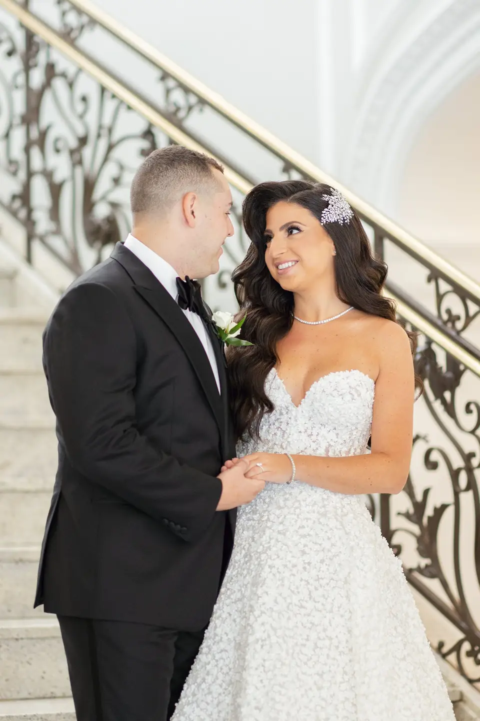 The bride and groom face each other at the foot of a marble staircase with a wrought-iron rail.
