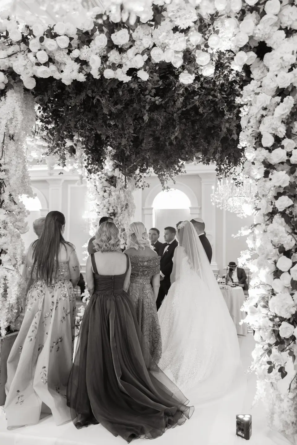 Seen from behind, guests beneath a canopy of white flowers at the ceremony. Black and white.
