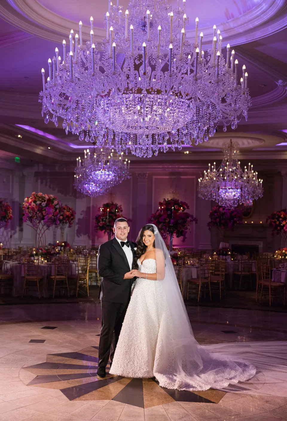The bride and groom in the ballroom beneath a great crystal chandelier, tables in bloom around them.