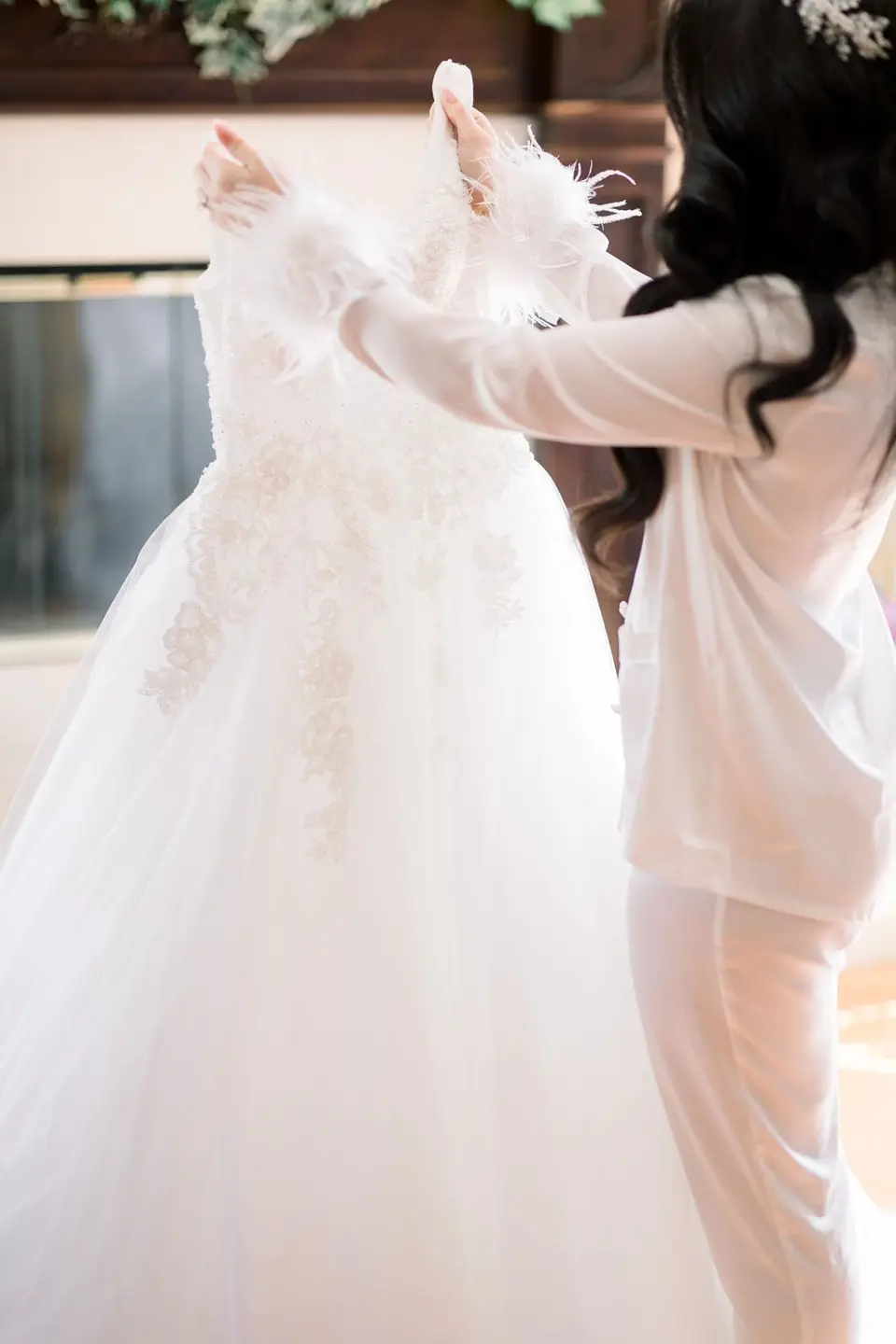 The bride in white pyjamas reaches for her gown as it is lifted from its hanger.