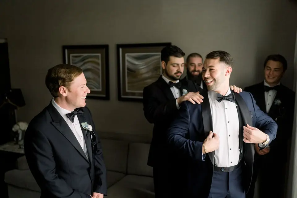 The groom's friends help him into his jacket as he laughs.