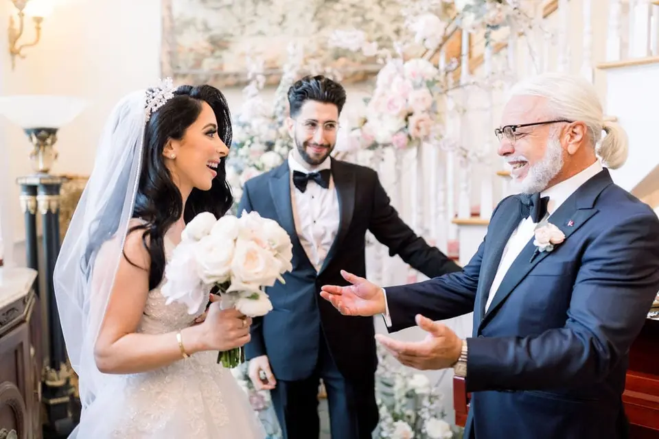 The bride's father greets her with open arms at the house, the bride holding white roses.