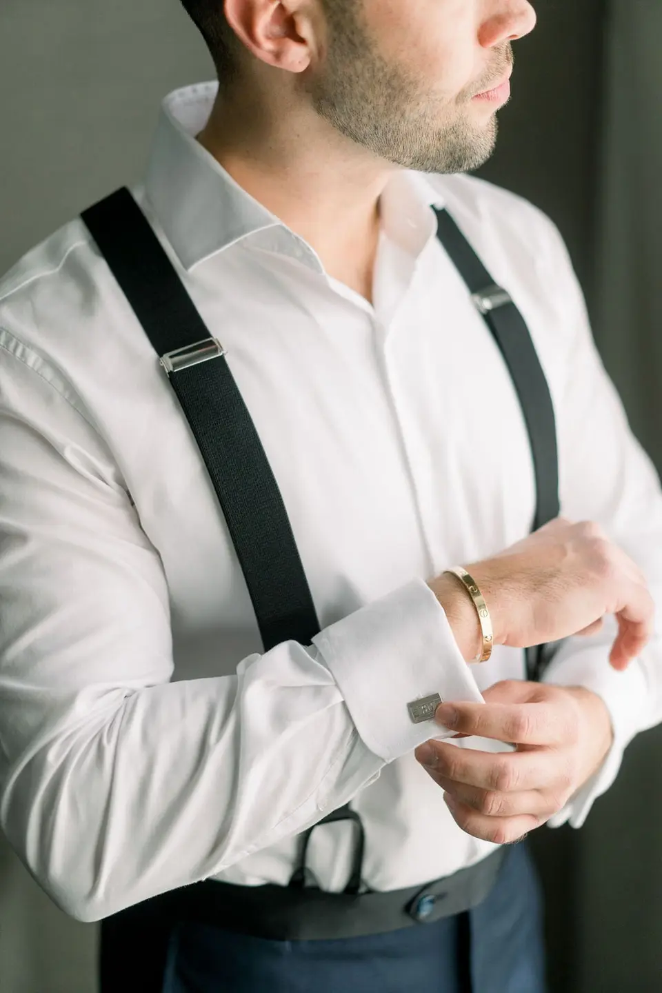 The groom in white shirt and suspenders fastens his cufflink.