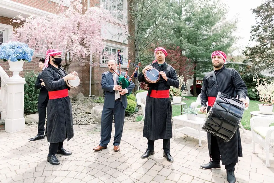 Musicians in black and red with drums and bagpipes play in the courtyard beneath a pink cherry tree.