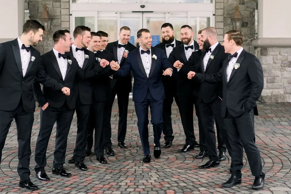 The groom laughs with his groomsmen in black tuxedos outside the house.