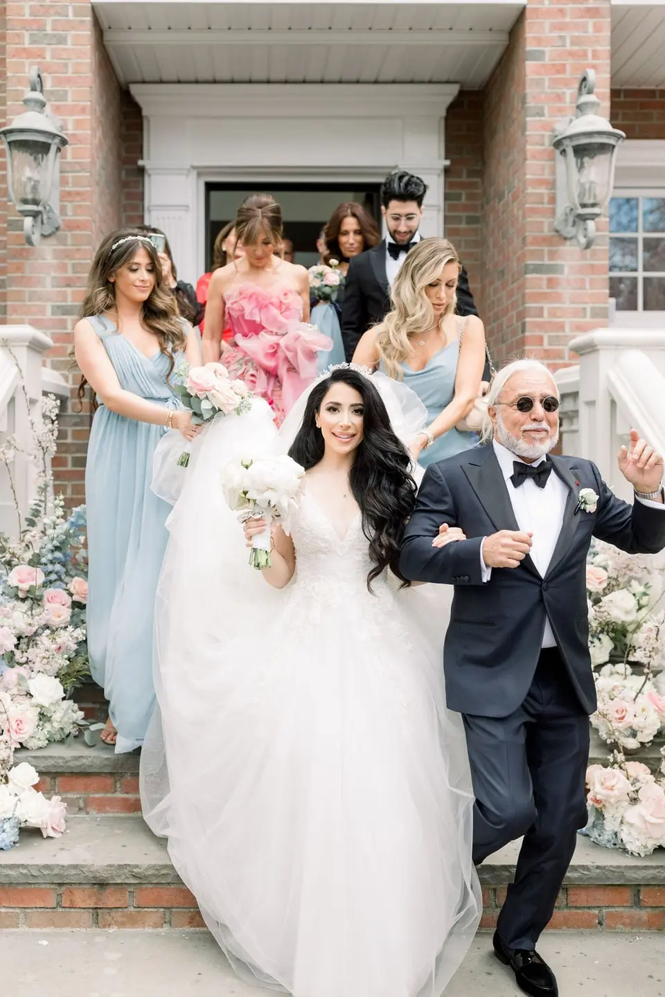 The bride comes down the front steps on her father's arm, the bridesmaids in pale blue behind them.