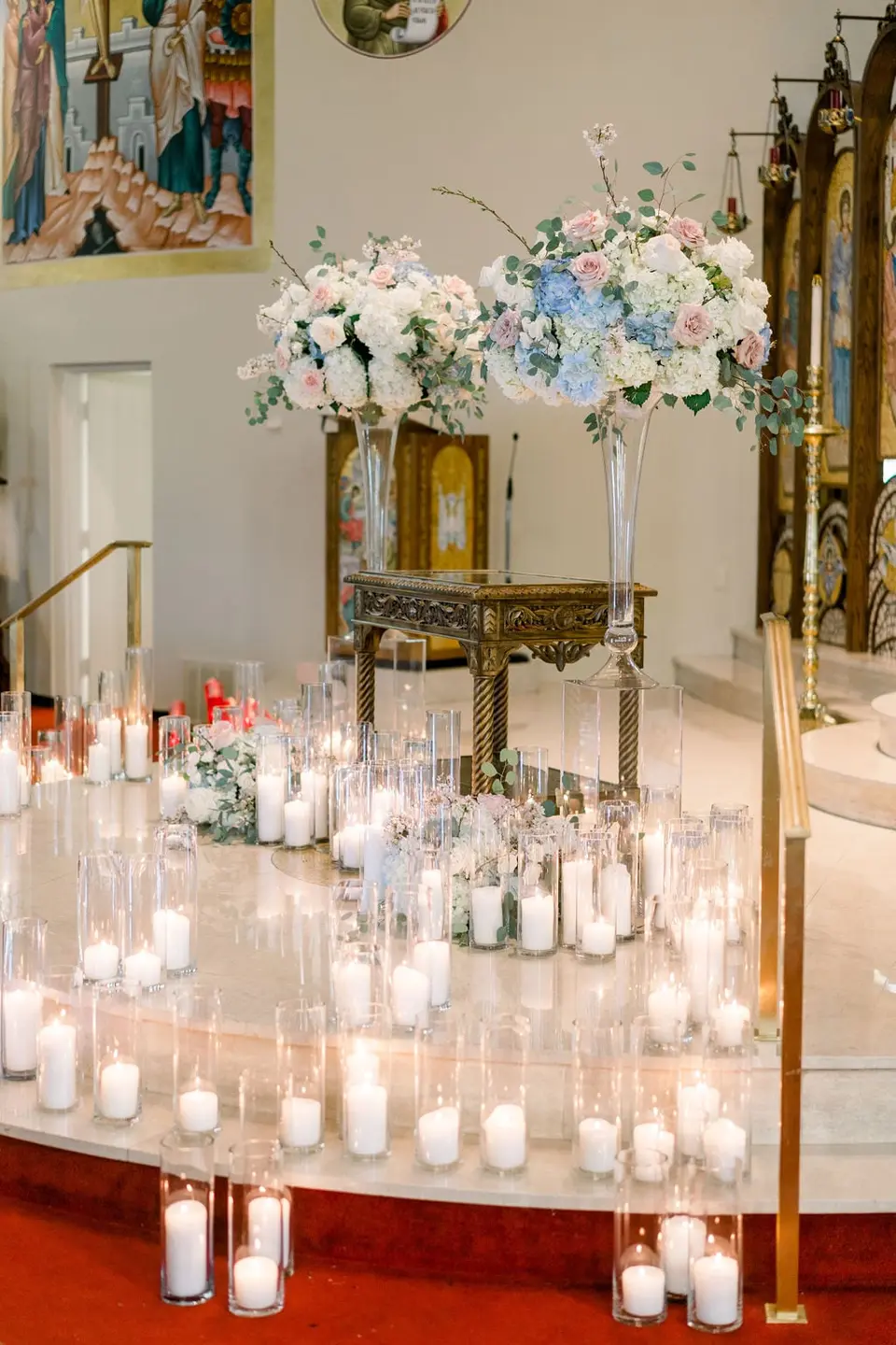 The altar steps lined with glass candle holders, tall arrangements of white and blue flowers beside them.