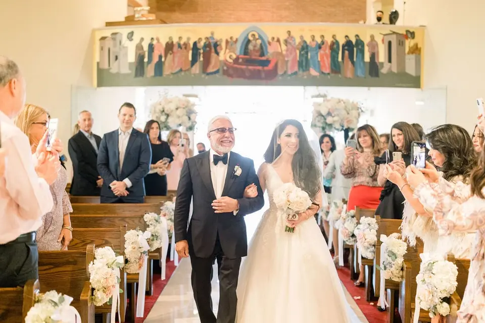 The bride walks down the aisle on her father's arm, guests turning to watch.