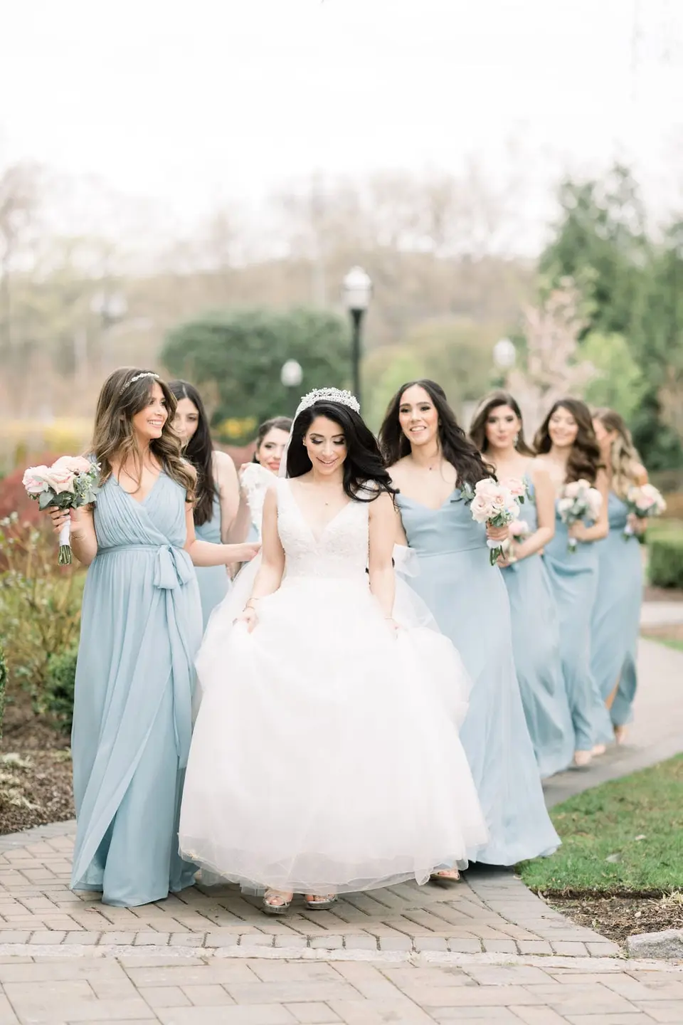 The bride walks with her bridesmaids in pale blue along a garden path.