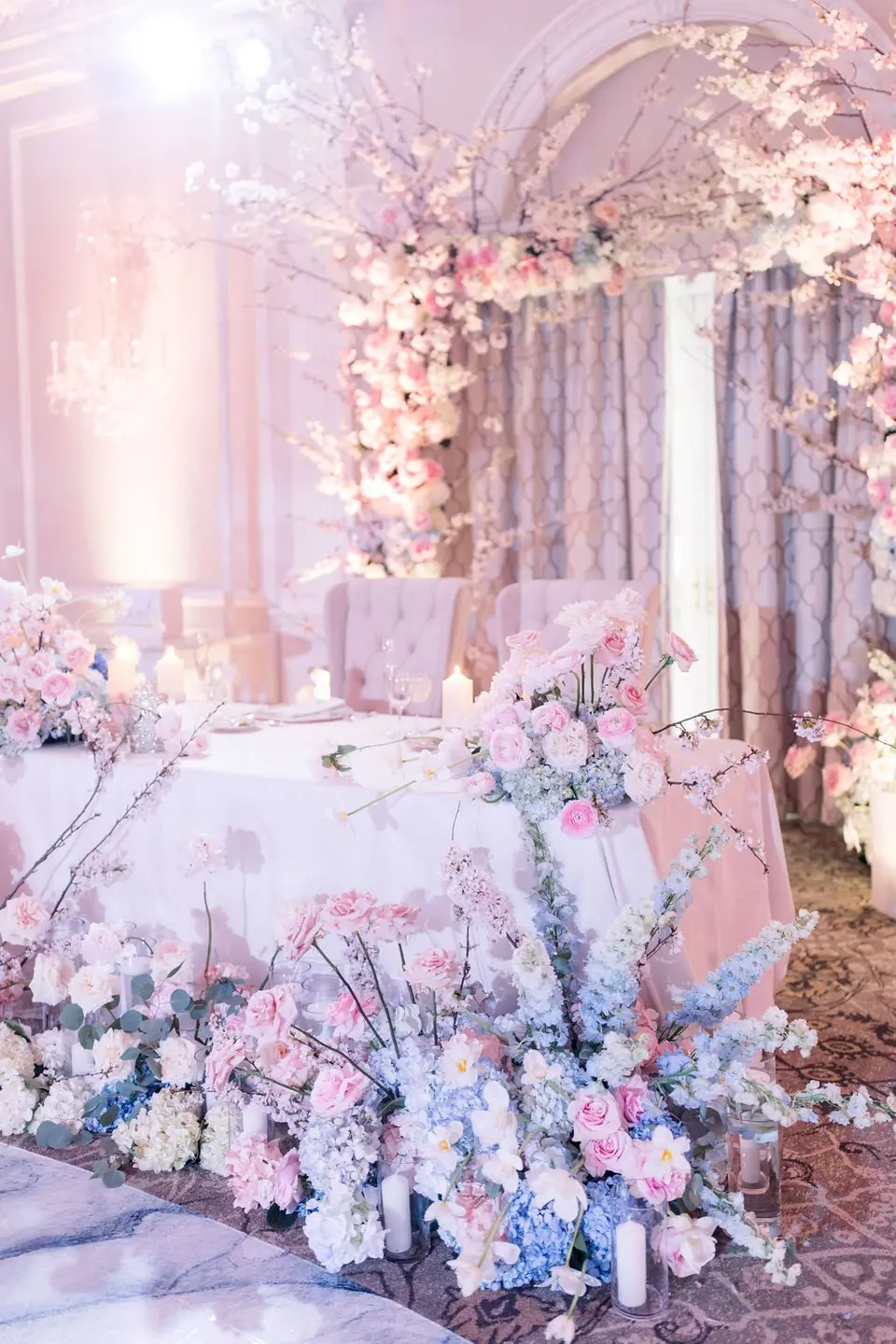 The sweetheart table framed by blossom branches, pink and blue flowers spilling to the floor.