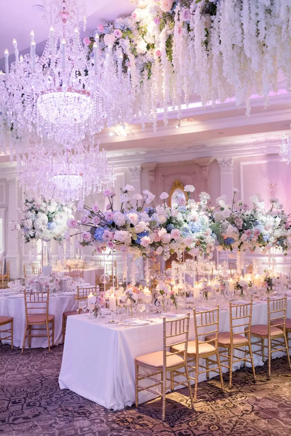 The ballroom beneath chandeliers and hanging white flowers, long tables with tall arrangements of pink and blue.