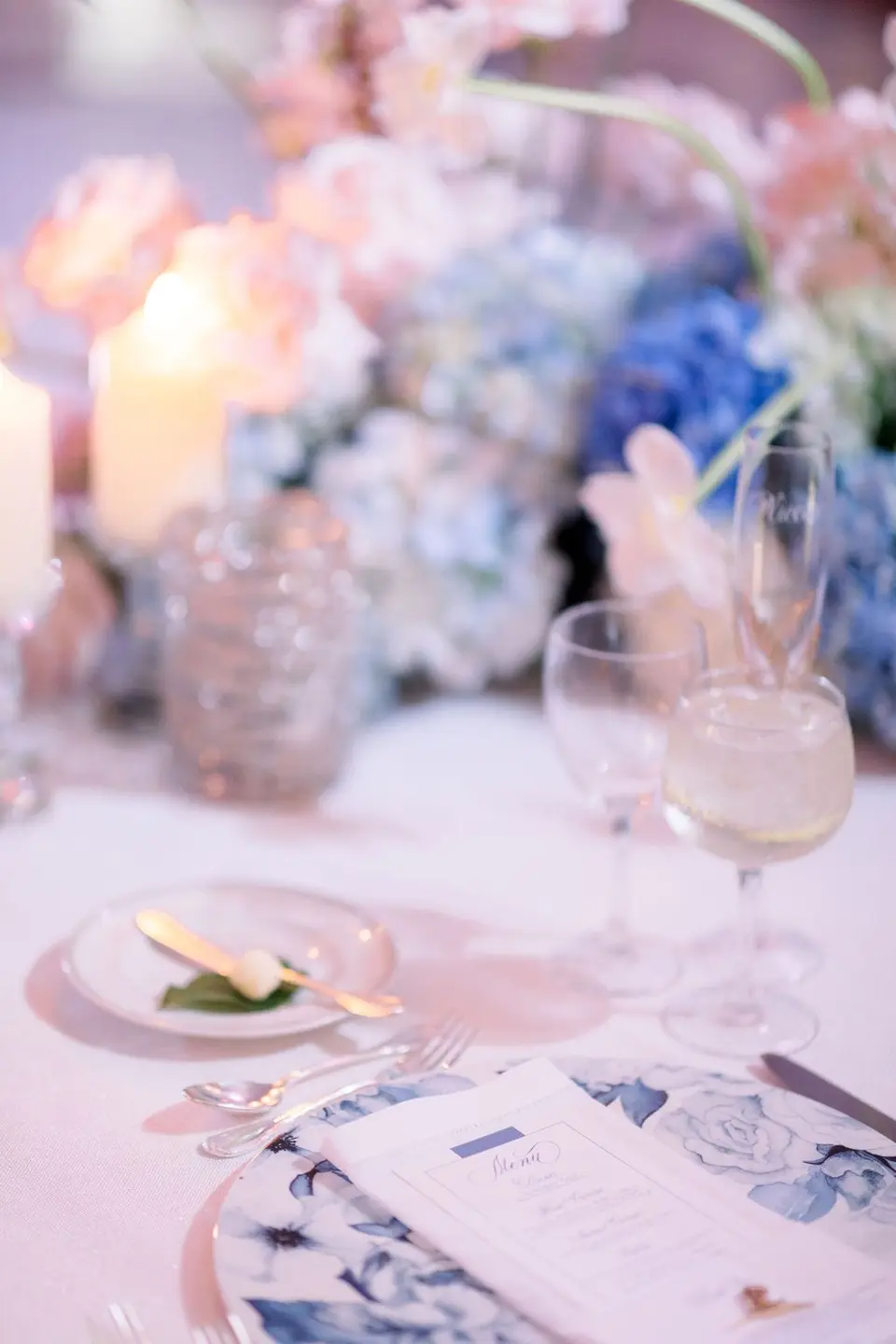 A place setting with a blue floral charger and a menu card, candles and blue hydrangeas behind.