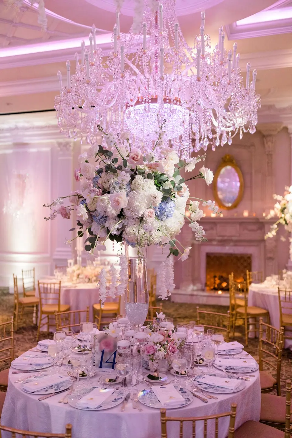 A round table beneath a crystal chandelier, a tall arrangement of white and blue flowers at its center.