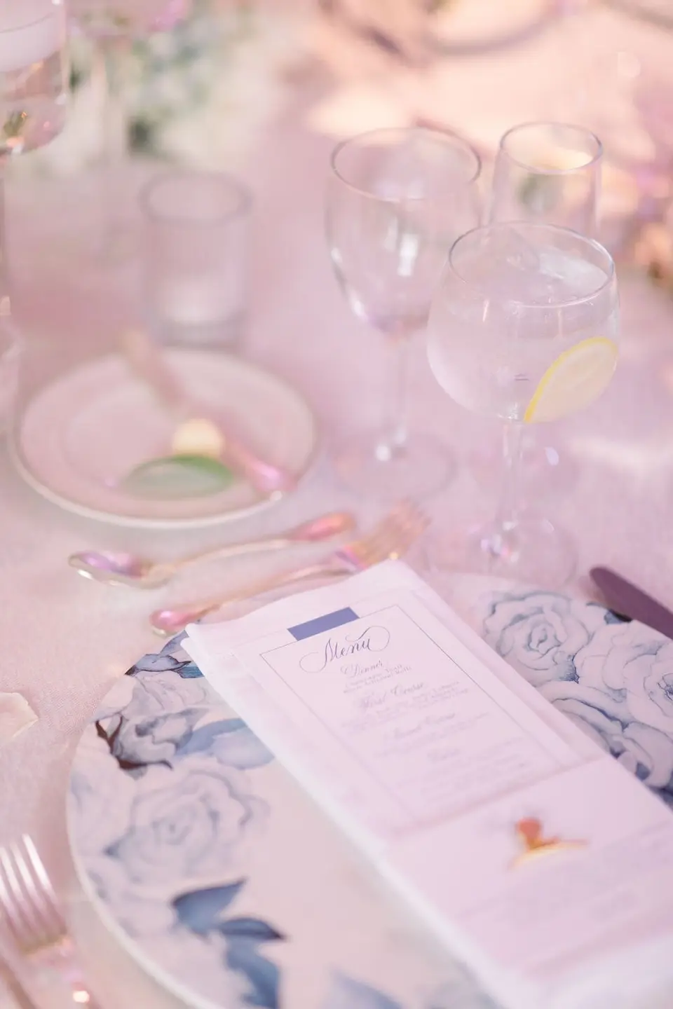 A menu on a blue and white floral charger, glasses catching the pink light.