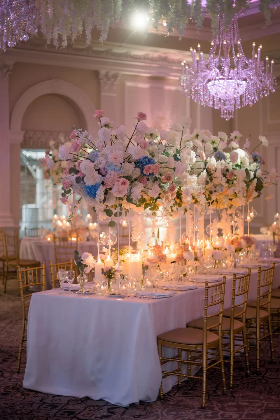 A long table glowing with candles beneath a tall arrangement of pink and blue flowers and a crystal chandelier.