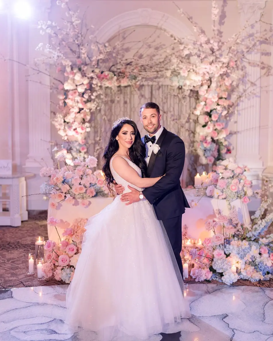 The bride and groom embrace in the ballroom before an arch of cherry blossom and blush roses, candles glowing on the marble floor.
