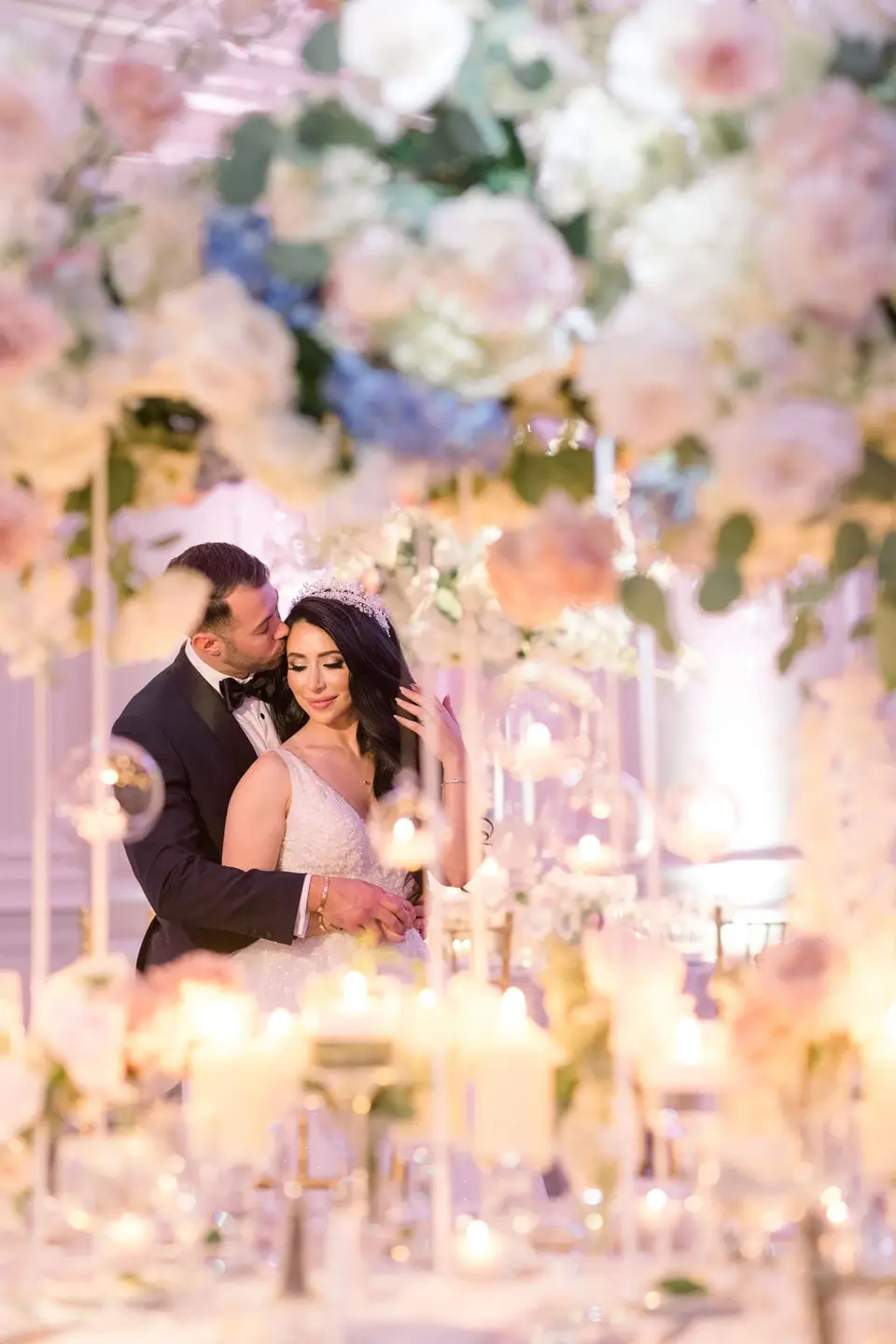 The groom kisses the bride's cheek, seen through candles and white flowers on the table.