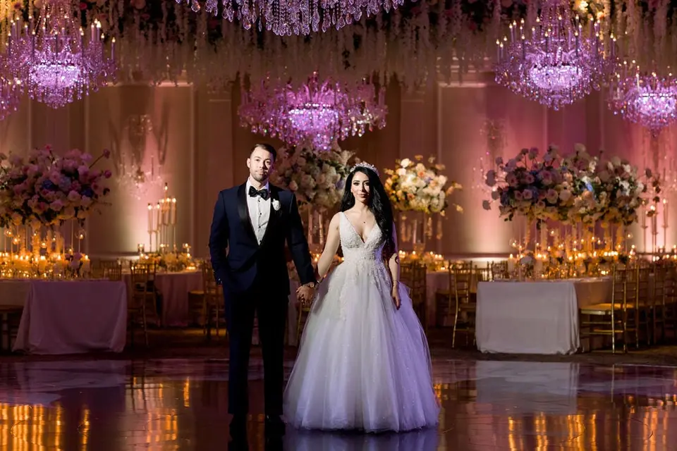 The bride and groom hold hands on the dance floor beneath pink-lit chandeliers.